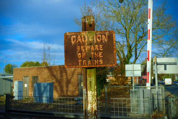 Warnschild an einem Bahnübergang-Zaun mit Bäumen, Strommasten, einem Gebäude, Containern, Fahrzeugen auf einer Straße und einem bewölktem Himmel.
