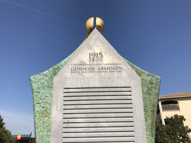 Ein steinernes Denkmal mit einer Uhr darauf, das mit Text beschriftet ist, steht in einem Park umgeben von Bäumen und Gebäuden unter einem klaren Himmel und erinnert an den 24. Jahrestag des Völkermords in Armenien.
