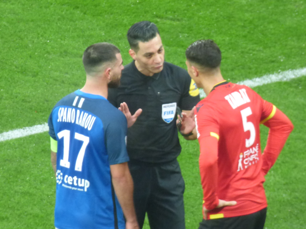Schiedsrichter in schwarzem Hemd und Hose, der mit zwei Männern in Brille auf einem Fußballfeld spricht; einer trägt ein blaues und rotes T-Shirt mit Text, der andere ein blaues und rotes T-Shirt.