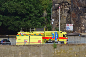Rettungswagen auf der Seite einer Straße neben einer Klippe geparkt, mit verschiedenen Straßenobjekten wie einer Wand, einem Geländer, Pfählen, Lampen und Texttafeln im Vordergrund und Bäumen und einem Auto im Hintergrund.