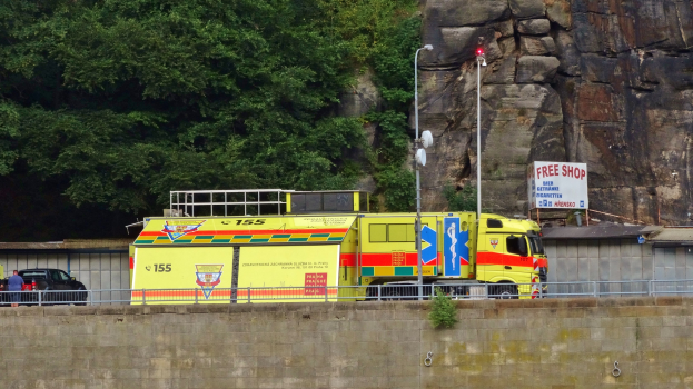 Rettungswagen auf der Seite einer Straße neben einer Klippe geparkt, mit verschiedenen Straßenobjekten wie einer Wand, einem Geländer, Pfählen, Lampen und Texttafeln im Vordergrund und Bäumen und einem Auto im Hintergrund.