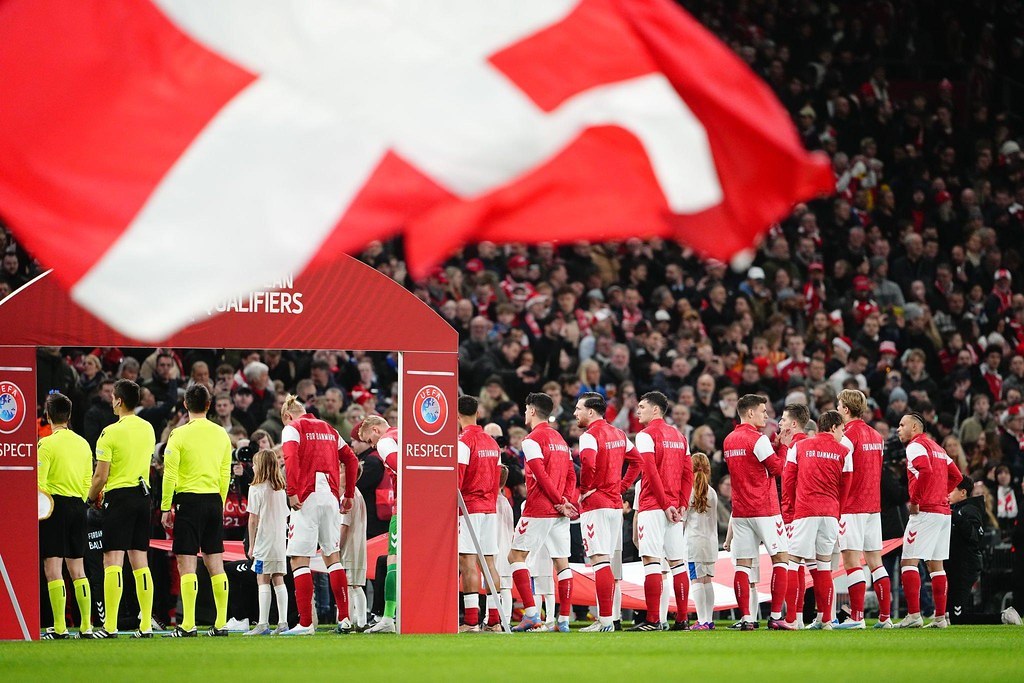 Eine Gruppe von Menschen steht auf einem Fußballfeld mit einer roten und weißen Flagge im Vordergrund, ein Bogen mit der Aufschrift "Bayern Munich vs Bayern Munich Betting Tips & Preview" im Hintergrund und eine große Menge im Stadion.