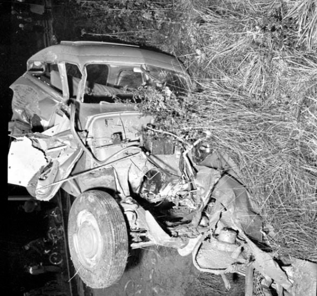 Ein Schwarz-Weiß-Foto eines stark beschädigten Autos am Straßenrand mit einer eingedrückten Front, zersplitterten Fenstern, einem Baum im Hintergrund und verstreuten Gegenständen rund um die Unfallstelle.