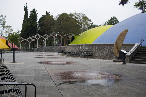 Ein Park mit einem zentralen Brunnen, umgeben von Bänken, Treppen mit Geländern, gewölbten Wänden, Zelten und Bäumen unter einem Himmel.
