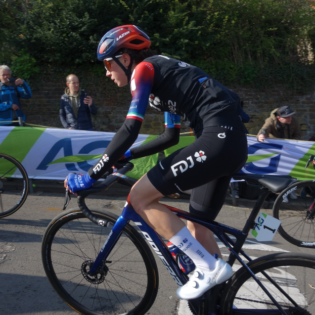 Professionelle Radrennfahrerin Katie Archibald trägt einen Helm, Schutzbrille, Handschuhe und Schuhe und fährt ein Fahrrad auf einer Straße neben einer Menschenmenge, mit einem Banner, einer Wand, Bäumen und einem Gebäude im Hintergrund.