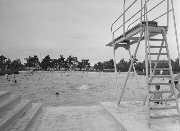Ein Schwarz-Weiß-Foto von Menschen, die im Wasser schwimmen, mit einem Lebensrettungsturm auf der rechten Seite mit Stufen und Bäumen, Pfählen und einem klaren Himmel im Hintergrund.