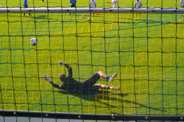Fussballspieler beim Abwehren eines Schusses mit einem Tor im Vordergrund und einigen Menschen und Bällen im Hintergrund.