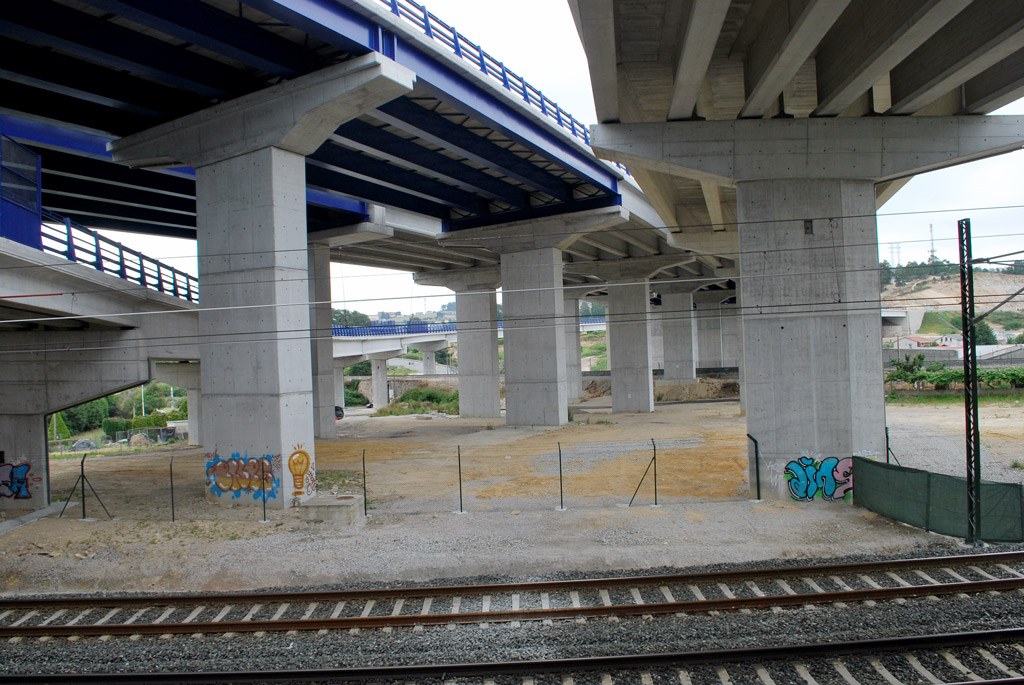 Eisenbahnschiene mit Graffiti unter einer Brücke, umgeben von Pfählen mit Drähten, Bäumen, Gebäuden und einem klaren blauen Himmel.