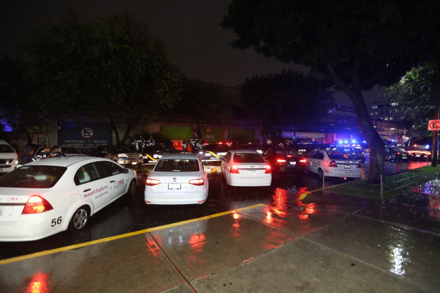 Polizeiautos auf einem Parkplatz bei Nacht mit einem Schild, Bäumen, Gebäuden, einer Fahne und einem bewölkten Himmel.
