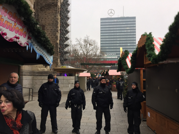 Polizisten in schwarzen Uniformen stehen vor einem geschmückten Weihnachtsmarkt in Berlin, Deutschland, mit festlichen Dekorationen, Gebäuden, Bäumen und einem klaren blauen Himmel im Hintergrund.