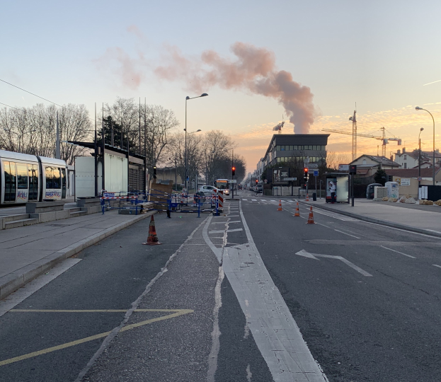 Straßenszene mit Verkehrskegeln, einem Bus, Laternenmasten, Bäumen, Gebäuden, Kränen und Rauch, der aus einem fernen Gebäude aufsteigt.