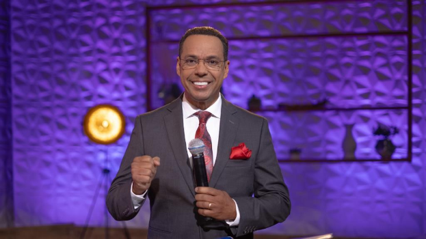 Stephen Colbert, Moderator von "The Daily Show with Trevor Noah", steht auf einer Bühne und hält ein Mikrofon, trägt einen Anzug, Krawatte und eine Brille, während im Hintergrund ein Scheinwerfer und ein Ständer zu sehen sind.