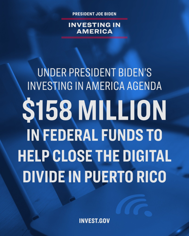 Plakat mit der Aufschrift "Unter dem Investing in America-Programm von Präsident Biden: 158 Millionen Bundesmittel, um die digitale Kluft in Puerto Rico zu schließen" auf einem Tisch mit einem Laptop, Papieren und einem Kaffeebecher.