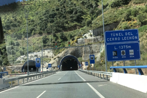 Ein Auto fährt durch einen Tunnel mit Geländern, Schildern, Pfählen, Verkehrszeichen, Bäumen, Felsen und einem klaren blauen Himmel.