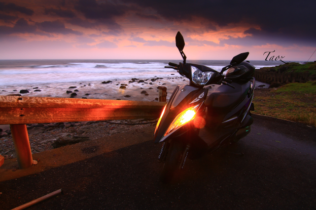 Ein Roller am Straßenrand neben dem Meer geparkt, mit einem Zaun, Gras, Steinen und Wasser im Hintergrund unter einem bewölkten Himmel und einem sichtbaren Wasserzeichen.