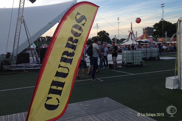 Eine belebte KarnevalsSzene mit mehreren Zelten, einem zentralen Mast, Menschen, Wagen auf der Straße, Bäumen, Strommasten und einem Eisenständer im Hintergrund unter einem Himmel.