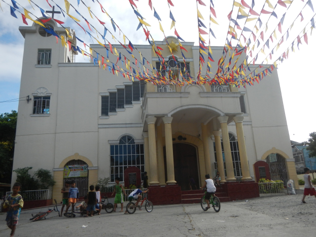 Eine Gruppe von Menschen, die vor einer weißen Kirche mit Fenstern, Säulen, Bögen, Türen und einem Kreuzsymbol Fahrrad fahren, mit Pflanzen, einem Zaun, einer Fahne, Bäumen und einem bewölkten Himmel im Hintergrund und bunten Fahnen, die an einem Seil oben befestigt sind.