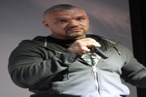 Ein Mann in einer schwarzen Kapuzenjacke und einem schwarzen Anzug mit einem weißen Hemd hält ein Mikrofon und steht selbstbewusst vor einer Wand.