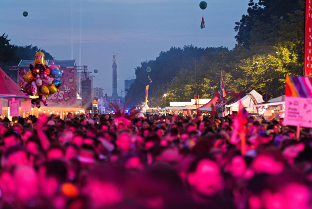 Eine große Menschenmenge auf einem nächtlichen Musikfestival mit farbigen Lichtern und Ballons, umgeben von Zelten, Bannern, Fahnen, Bäumen, einem Turm und einem sternenklaren Himmel.
