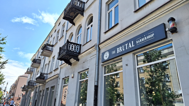 Brutalistische Barbershop-Fassaden in Berlin mit Betongebäuden, Fenstern, Geländern, Balkonen und Namensschildern, Bäumen, Laternenmasten, Fußgängern und einem bewölkten Himmel.