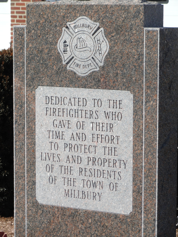 Denkmstein mit einer Plakette für Feuerwehrleute in Millbury, vor einem Backsteingebäude mit umgebender Vegetation.