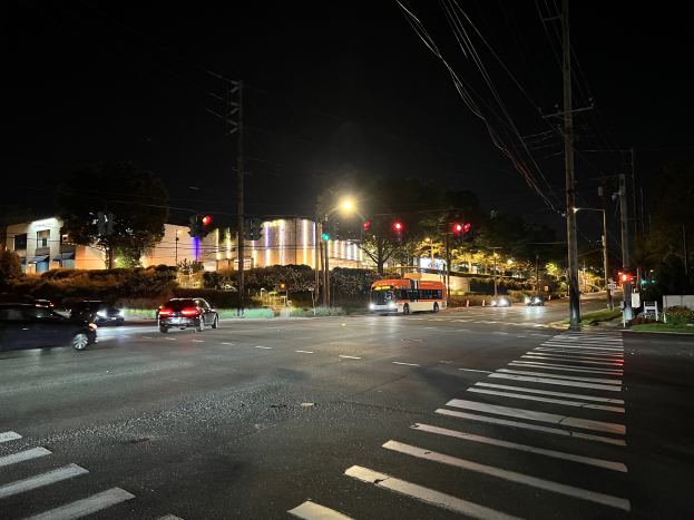 Eine nächtliche Stadtstraße mit fahrenden Autos und einem Bus, beleuchtet von Straßen- und Verkehrsschildern, gesäumt von Gebäuden, Bäumen und Gras, unter einem dunklen Himmel.