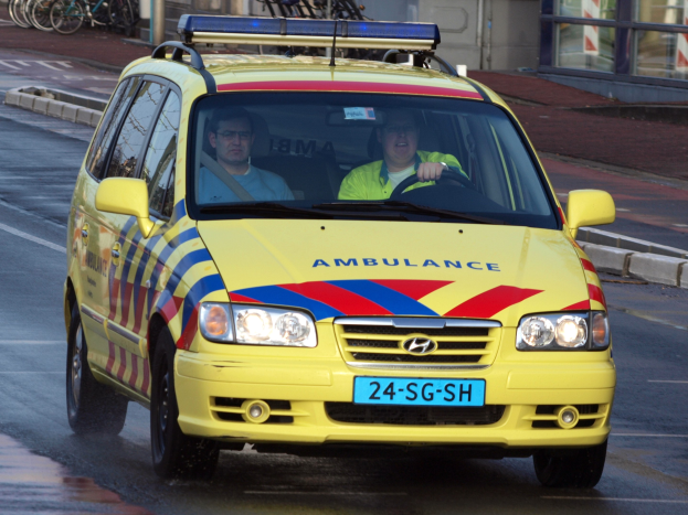 Ein gelber Krankenwagen mit zwei sichtbaren Insassen fährt an parkenden Fahrrädern auf einem Gehweg vorbei, mit einem Glaswandgebäude im Hintergrund.