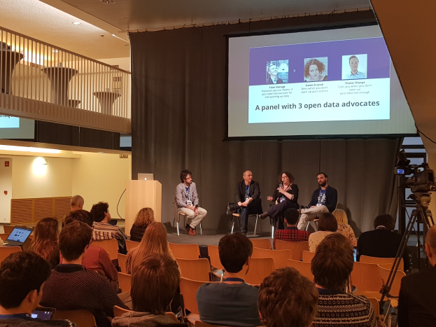 Eine Gruppe von Menschen sitzt auf Stühlen und schaut auf einen Projektionsschirm, auf dem ein Laptop auf einem Tisch steht, eine Kamera auf einem Stativ rechts daneben und Geländer, Lampen und eine Wand im Hintergrund während einer Podiumsdiskussion über Open-Data-Aktivisten.