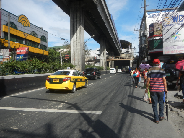 Ein gelbes Taxi fährt auf einer Stadtstraße mit hohen Gebäuden, Passanten auf dem Bürgersteig mit Regenschirmen, einer Brücke im Hintergrund, bewölktem Himmel und Strommasten mit Überlandleitungen.