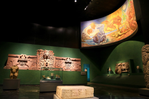Ein Museumsraum mit verschiedenen Artefakten, darunter Skulpturen, Gegenstände auf Tischen, eine Texttafel, eine Wandmalerei und Deckenleuchten im Anthropologischen Museum von Mexiko-Stadt.