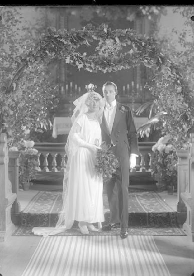 Schwarz-weißes Foto eines Mannes und einer Frau, die unter einem Blumenschmuckbogen stehen, die Frau hält einen Strauß.