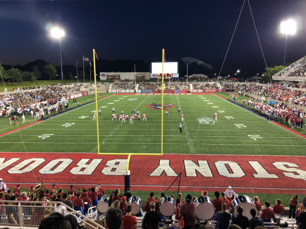 Ein nächtliches Fußballspiel in einem Stadion mit Spielern auf dem Feld, Zuschauern auf Stühlen, Musikern, einem Metallzaun, Laternen, Fahnen, einem Bildschirm, Bäumen, Hügeln und einem sternenklaren Himmel.