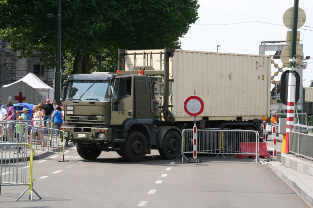Lkw am Straßenrand mit einer Gruppe von Menschen auf dem Gehweg, umgeben von Absperrungen, Schildern, Straßeninfrastruktur und Gebäuden unter einem klaren blauen Himmel.