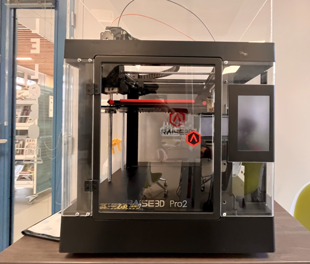 Ein 3D-Drucker auf einem Holztisch mit einer Glastür links und einem Stuhl rechts, vor Regalen mit Gegenständen im Hintergrund.