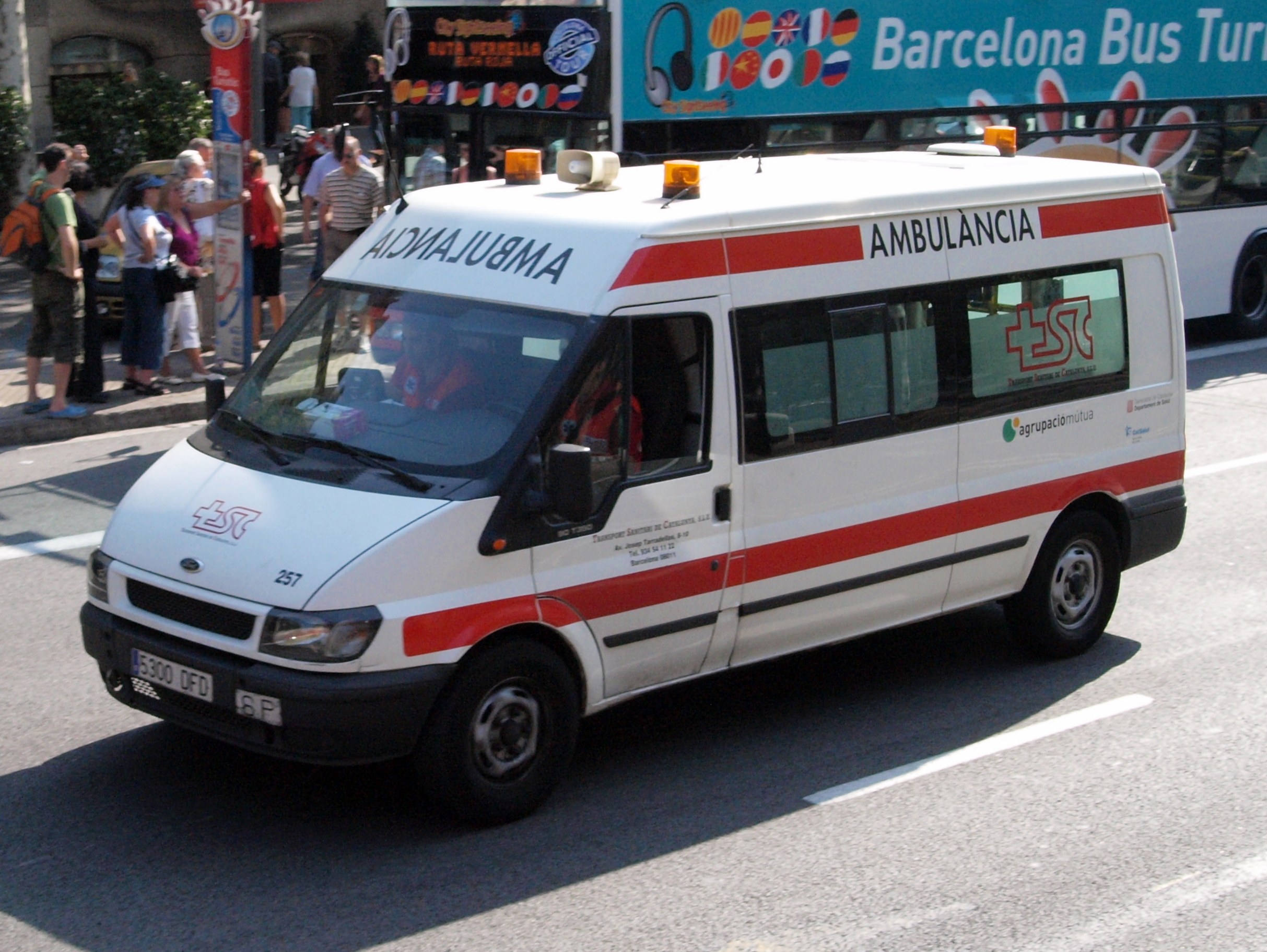 Rettungswagen neben einem Bus auf einer Straße fahrend, mit Passanten auf dem Gehweg und Gebäuden, Bäumen und einem klaren Himmel im Hintergrund.