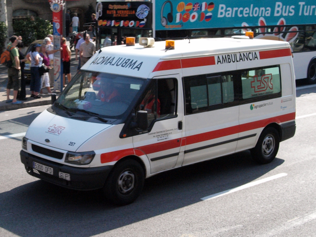 Rettungswagen neben einem Bus auf einer Straße fahrend, mit Passanten auf dem Gehweg und Gebäuden, Bäumen und einem klaren Himmel im Hintergrund.