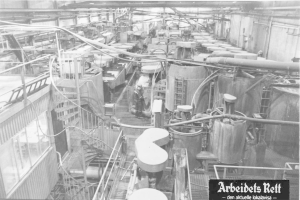 Schwarzes und weißes Foto eines großen Fabrikinneren mit einer Person, die zwischen Industriegeräten, Rohren und konstruktiven Elementen steht; Text unten: "Arbeidets Rett, die größte Fabrik der Welt."