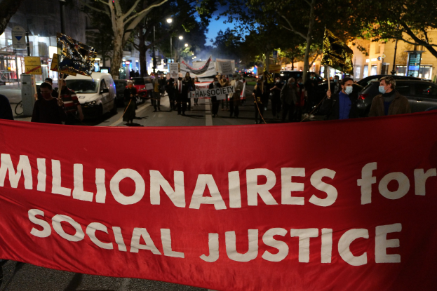 Eine Gruppe von Menschen marschiert mit einer roten Fahne, auf der 'Millionäre für soziale Gerechtigkeit' steht, eine Straße entlang, mit Bäumen, Gebäuden und vorbeifahrenden Fahrzeugen im Hintergrund.