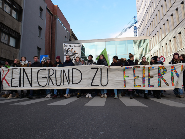 Protestierende auf einer Straße halten ein Transparent mit der Aufschrift "Kein Grund zu Feiern" gegen die deutschen Sparmaßnahmen, mit Gebäuden, einem Schild, einem weiteren Transparent mit Text, einem Kran und einem bewölkten Himmel im Hintergrund.