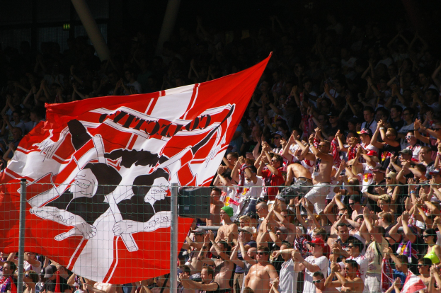 Große Menschenmenge in einem Stadion mit einer rot-weißen Fahne, Maschendrahtzaun im Vordergrund und Säulen vor einem dunklen Hintergrund.