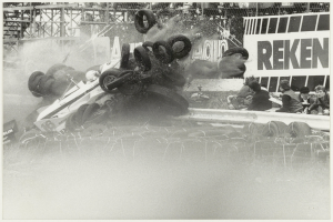 Ein Schwarz-Weiß-Foto eines verunglückten Rennwagens, umgeben von Reifen, mit einer Gruppe von Menschen rechts und einer Tafel mit Text im Hintergrund.