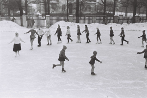 Schwarz-weißes Foto von Kindern, die auf einer schneebedeckten Eisbahn Schlittschuh laufen, umgeben von einem Zaun, Bäumen und Gebäuden.