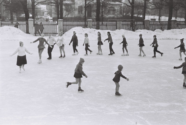 Schwarz-weißes Foto von Kindern, die auf einer schneebedeckten Eisbahn Schlittschuh laufen, umgeben von einem Zaun, Bäumen und Gebäuden.