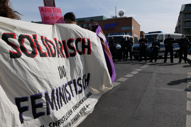 Gruppe von Menschen marschiert auf einer Straße und hält ein 'Solidarität und Feminismus'-Schild, mit geparkten Fahrzeugen, Gebäuden, einer Schüsselantenne und einem klaren blauen Himmel im Hintergrund.