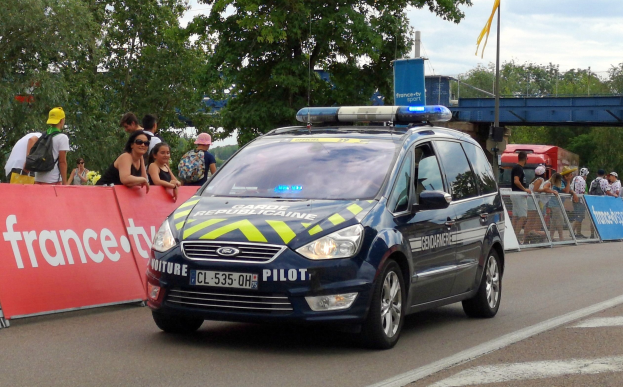 Polizeifahrzeug fährt an einer Menge mit Schildern, Geländern, Bäumen, einer Brücke, einer Flagge und einem bewölkten Himmel im Hintergrund vorbei.