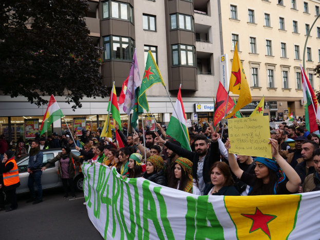 Eine große Gruppe von Menschen marschiert auf einer Straße mit Fahnen und Transparenten, begleitet von einem geparkten Auto und einem Baum auf der linken Seite, mit Gebäuden und Namensschildern im Hintergrund.