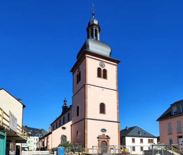 Große rosafarbene Kirche mit einem zentralen Uhrenturm, umgeben von einem Metallzaun, vor einem bewölkten Himmel, mit benachbarten Gebäuden, Fahrzeugen, Pflanzen, einem Schild und einer Fahne.