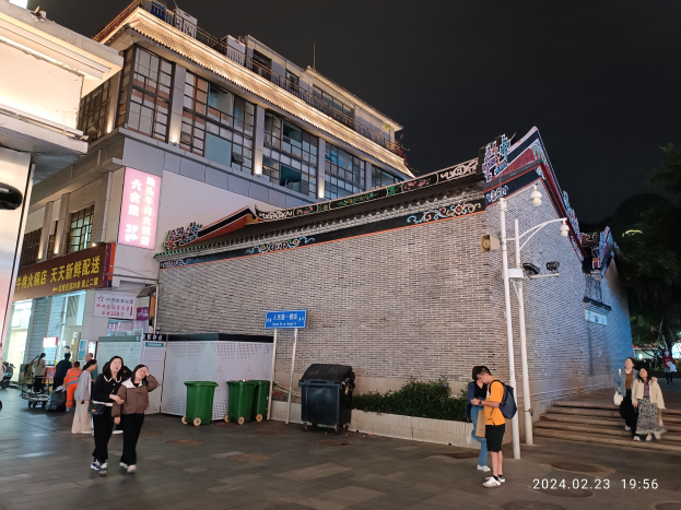 Eine Gruppe von Menschen, die eine nachtlich beleuchtete Straße mit Gebäuden, Schildern, Pflanzen und Mülltonnen entlanggehen, einige tragen Taschen.