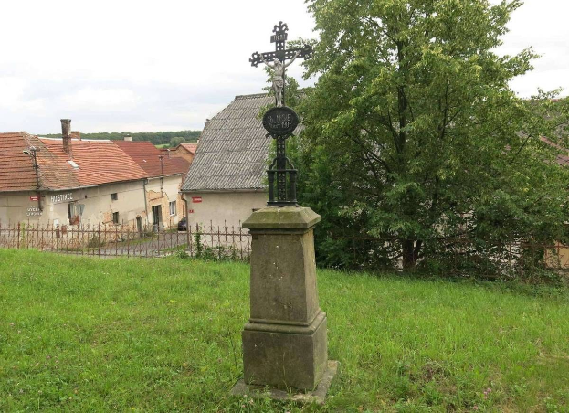Ein großes Kreuz steht als Holocaust-Mahnmal in einem von einem Metallzaun umgebenen Grasfeld, mit Bäumen, Häusern und einem bewölkten Himmel im Hintergrund.