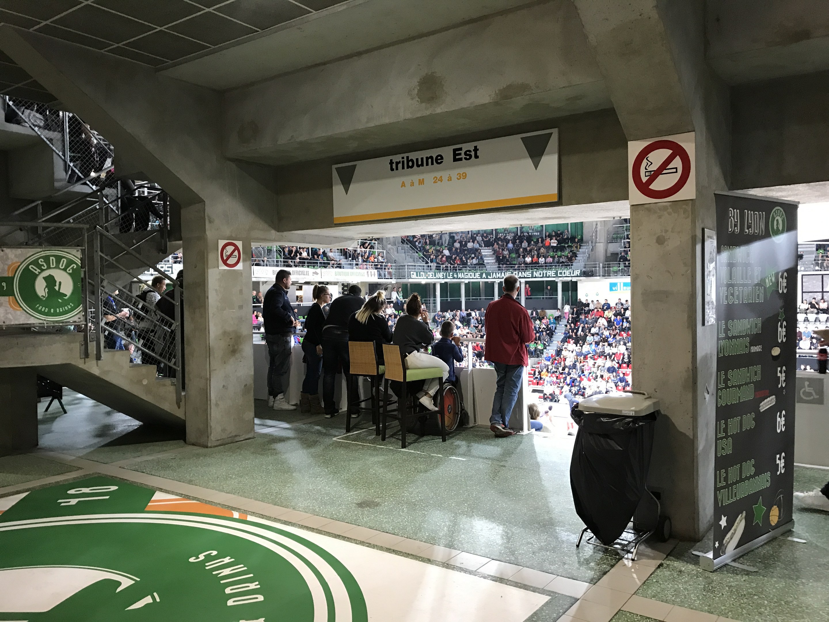Eine Gruppe von Menschen steht vor einem sitzenden Publikum in einem Stadion während eines Boston Celtics vs New York Jets-Events, mit Schildern, einem Müllcontainer, einer Tafel mit Text, einer Treppe mit Geländern und Deckenleuchten.